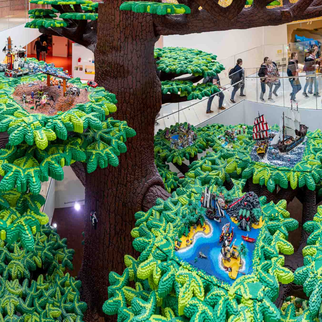 This is a massive lego sculpture found in lego headquarters. It's a tree that spans at least 3 stories of the open lobby area. The branches of the tree each have an intricate lego scene built on them. In the background, there are people standing on a balcony viewing the structure. The people are small, showing the incredible scale of the sculpture.